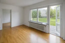 Tom lägenhet med ljust trägolv, vita väggar och öppen balkongdörr mot en grön gård.