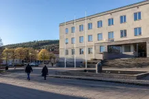 Funkisbyggnad med flaggstänger och bred trappa vid ett stenlagt torg där två personer går förbi.