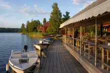 Uteservering på en brygga vid sjön med förtöjda båtar och röda trähus i kvällssol.