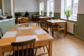 Matsal med gröna träpaneler, dukade träbord med glas och krukväxter i fönstren i dagsljus.