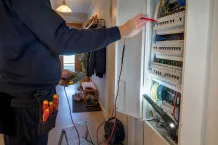 Elektriker mäter i en öppen elcentral i en hall, med skruvmejslar i bältet.
