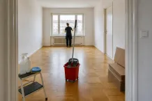 Tömd lägenhet med parkettgolv där en städare arbetar vid fönstret och mopp, hink och rengöringsmedel står framme.