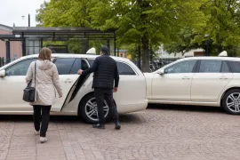 Två vita bilar vid en busskur under lövträd där en resenär med väska går mot den öppnade bakdörren.